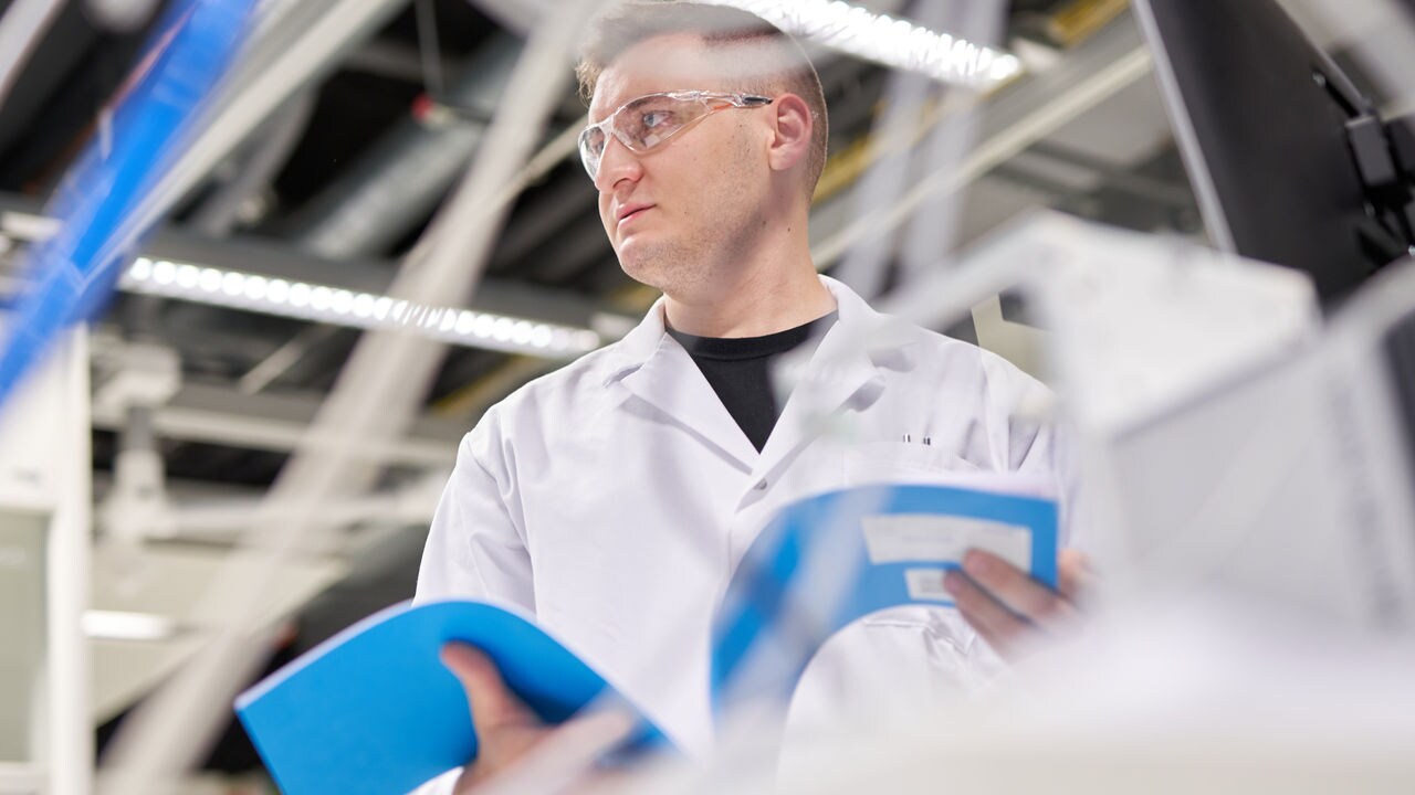 A scientist in a white lab coat with transparent glasses holds a notebook.